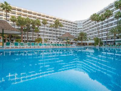 Großes Hotel mit Pool, Sonnenliegen und Palmen unter klarem Himmel.