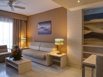 Soggiorno d'hotel con divano beige, poltrona, tavolo e quadro, luce naturale e lampade.