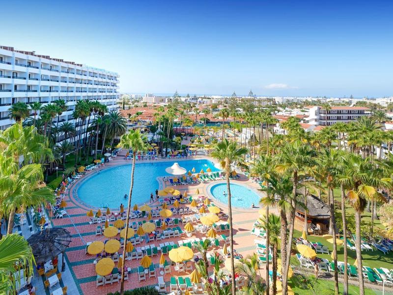 Vista di un hotel con piscina circondata da palme e lettini gialli sotto un cielo sereno.