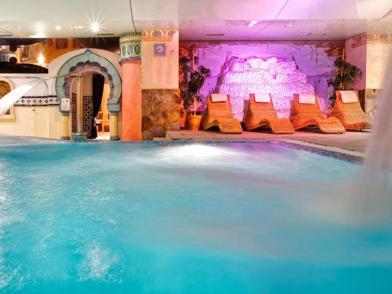 Piscina interna con getti d'acqua, lettini e illuminazione viola soffusa.