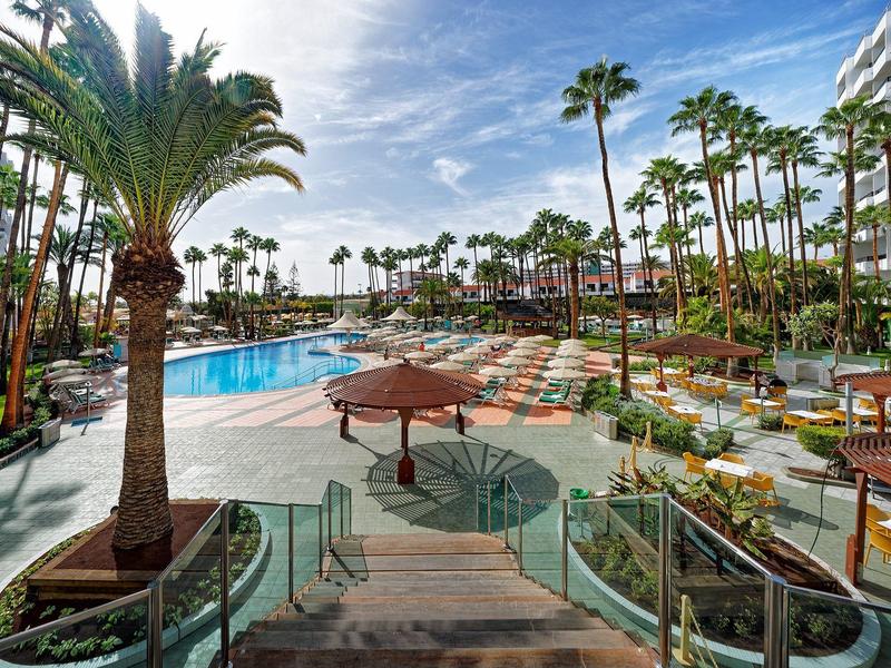 Piscina circondata da palme e lettini sotto un cielo azzurro in un hotel.