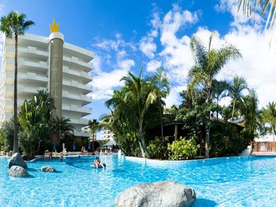 Großer blauer Pool mit Palmen und weißem Hotelgebäude unter blauem Himmel und Wolken.