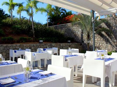 Terrassenrestaurant mit weißen Tischen und Stühlen, blauen Tischdecken und Palmen im Hintergrund.