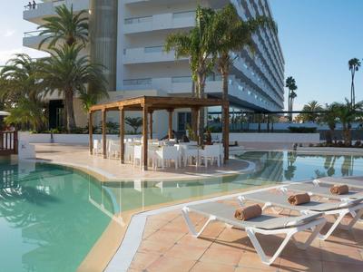 Hotelpool mit weißen Liegen, Palmen und einem überdachten Sitzbereich bei klarem Himmel.