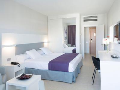 Helles Hotelzimmer mit Doppelbett, lila Bettdecke, Schreibtisch, Stuhl und Flachbildfernseher an weißer Wand.