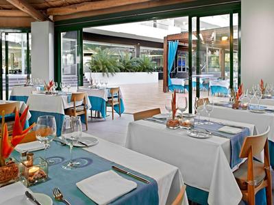 Helles Restaurant mit Tischen, weiß-blauen Tischdecken und modernen Holzstühlen. Große Fenster.