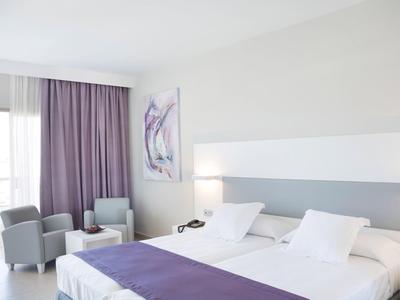 Helles Hotelzimmer mit zwei Betten, violetten Vorhängen, Sesseln und hellem Wandbild.