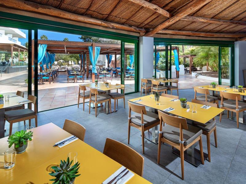 Lichte eetruimte met gele tafelkleden en houten stoelen met uitzicht op zitplaatsen buiten.