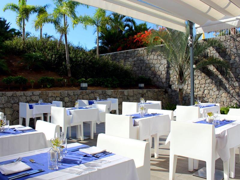 Buiten eetgedeelte van hotel met witte tafels en stoelen onder een luifel, omgeven door palmbomen.