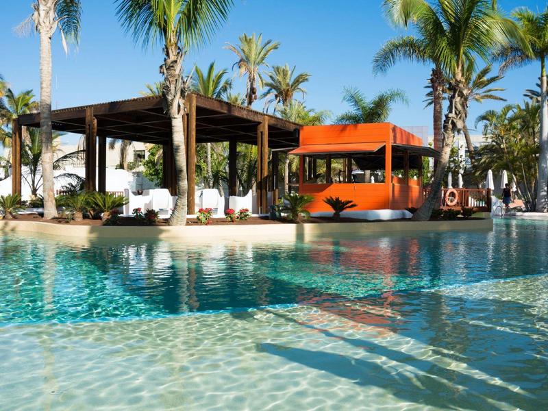 Buitenzwembad met palmbomen, overdekte zithoek en fel oranje zitplaatsen onder een heldere blauwe lucht.