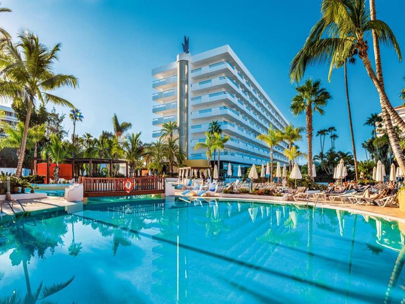 Zonnig tropisch hotel met blauw zwembad, palmbomen, ligstoelen en heldere lucht.