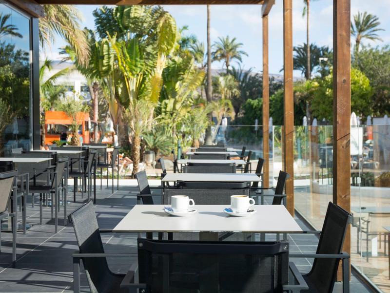 Buitenterras van hotel met tafels, stoelen en tropische planten bij zonnig weer.