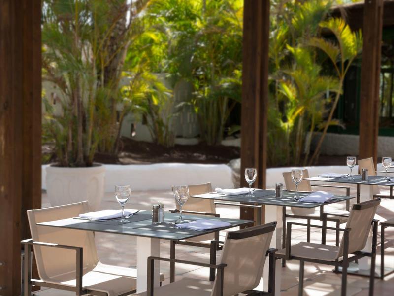 Buiten eetgedeelte met tafels en stoelen onder een schaduwrijke patio omgeven door groen.