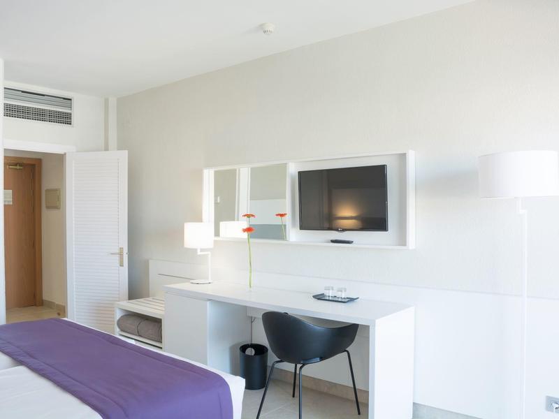 Lichte hotelkamer met wit meubilair, bureau, zwarte stoel en aan de muur gemonteerde tv