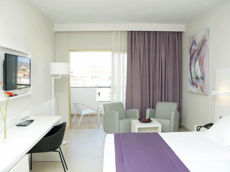 Modern hotelkamer met paarse bedloper, twee fauteuils, bureau en groot raam met gordijnen.