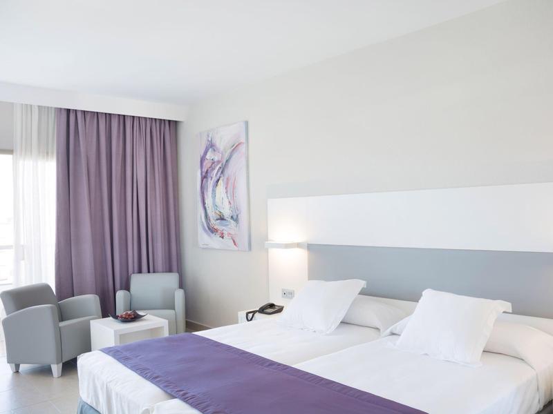 Lichte hotelkamer met twee aparte bedden, paarse accenten, fauteuils en groot raam met gordijnen.