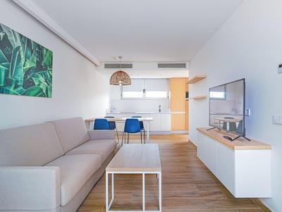 Nowoczesny salon z szarą sofą, telewizorem i otwartą kuchnią w jasnym apartamencie.