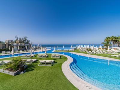 Moderner Hotelpool mit Liegestühlen, grüner Rasenfläche und Blick auf das Meer unter klarem blauem Himmel.