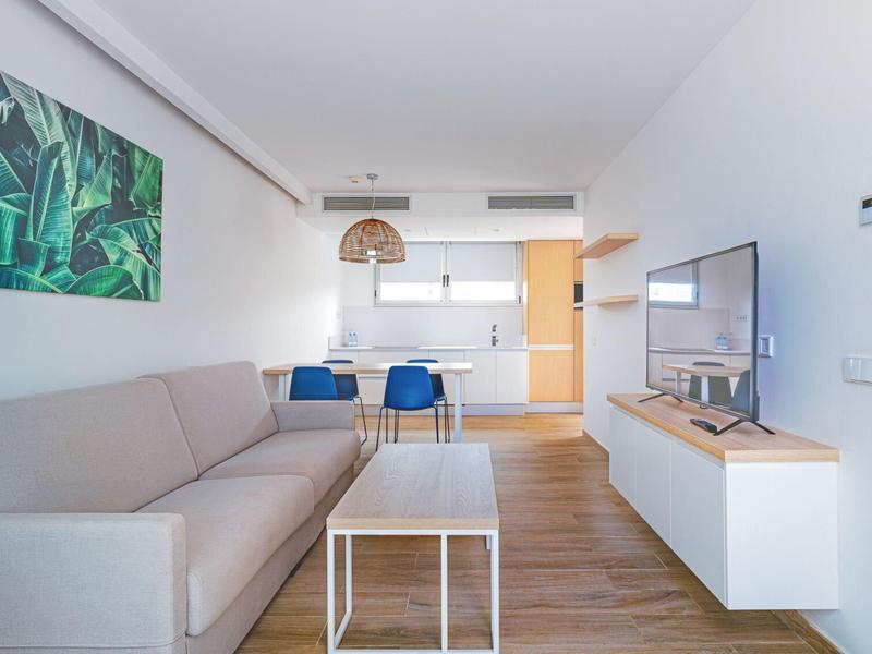 Nowoczesny salon z szarą sofą, telewizorem i otwartą kuchnią w jasnym apartamencie.