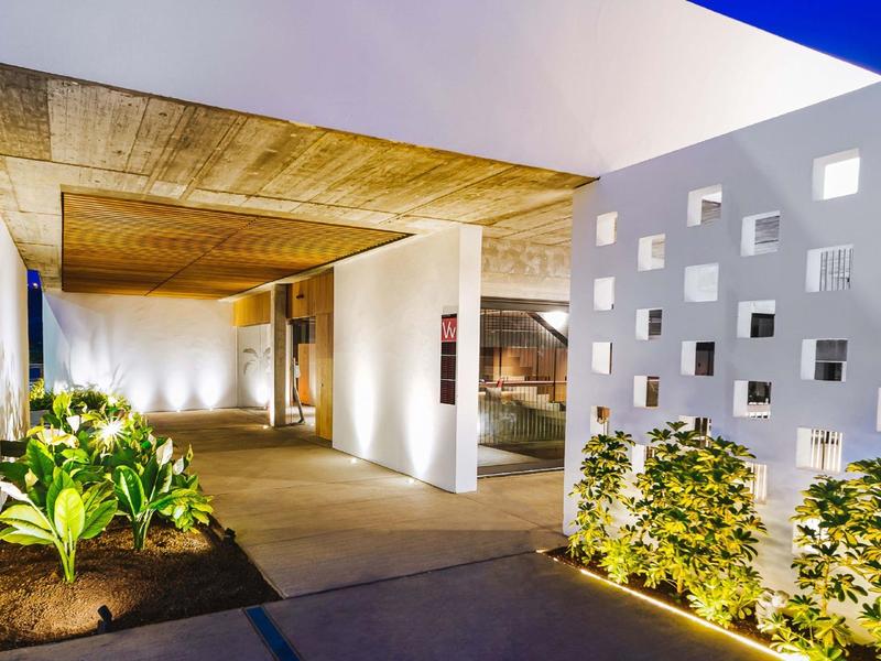 Nowoczesny korytarz hotelowy z oświetleniem, roślinami i białą ścianą z kwadratowymi otworami.