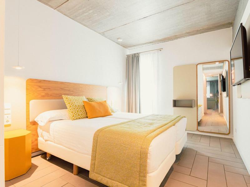 Jasny pokój hotelowy z podwójnym łóżkiem, żółtą pościelą, dużym lustrem i telewizorem na ścianie.