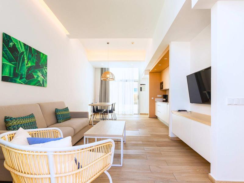 Jasny pokój hotelowy z sofą, fotelem, stolikiem i telewizorem, nowoczesne wnętrze z drewnianą podłogą.