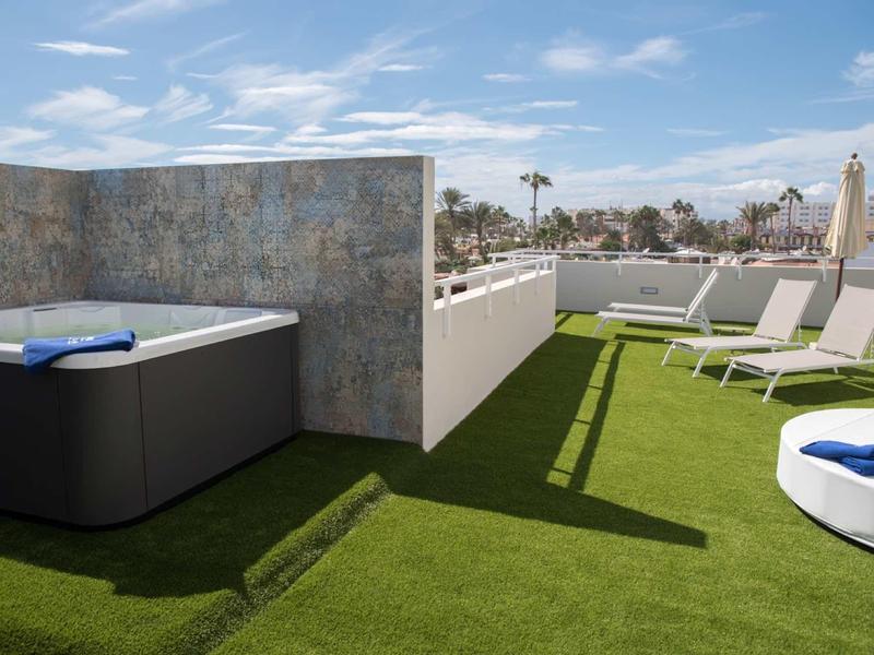 Rooftop terrace with spa area and lounge chairs on green artificial grass under sunny sky.