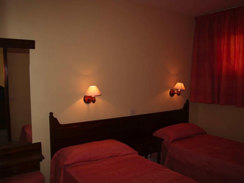 Zimmer mit zwei Einzelbetten, dunklem Holzkopfteil, roten Decken und warmem Wandlicht.