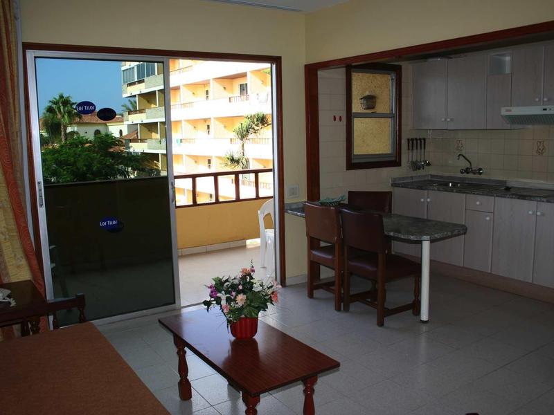 Wohnzimmer mit Holztisch, Stühlen, kleiner Küchenzeile und Balkon mit Aussicht auf Gebäude.