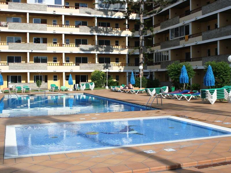 Mehrstöckiges Hotel mit blauen Sonnenschirmen, Liegestühlen und zwei Schwimmbecken im Vordergrund.