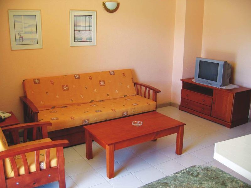 Kleines Wohnzimmer mit orangem Sofa, Holzstuhl, Tisch, TV auf Holzkommode und zwei Bildern an der Wand.