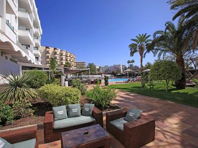 Terrasse d'hôtel avec sièges, jardin, palmiers et vue sur la piscine sous un ciel bleu.