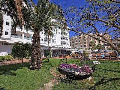 Green garden with palm trees and hanging chair in front of a white hotel building under blue sky.