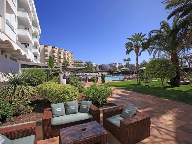 Hotel garden with lounge furniture and swimming pool, surrounded by palm trees and buildings.
