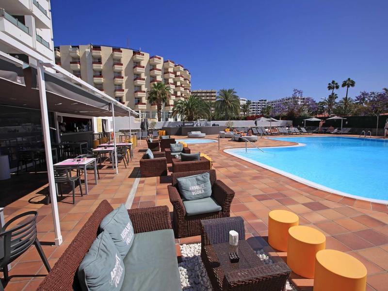 Hotel pool area with comfortable seating and sun umbrellas under a blue sky.