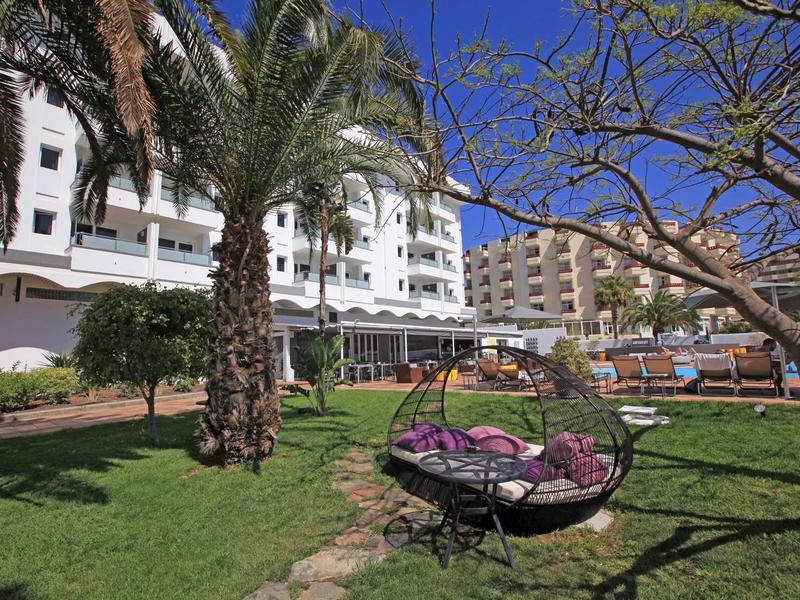 Green garden with palm trees and hanging chair in front of a white hotel building under blue sky.