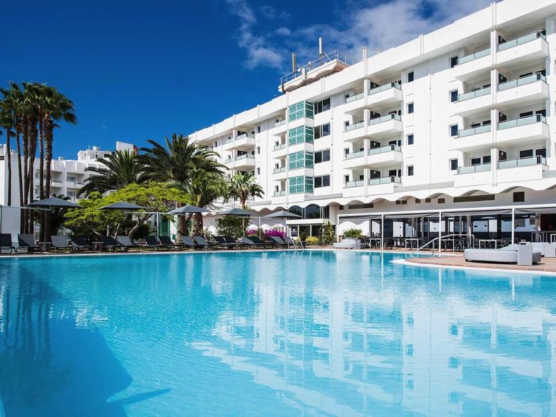 Modern hotel with large outdoor pool, palm trees, and umbrellas under blue sky.