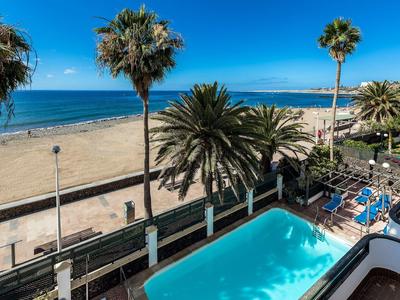 Widok na plażę z basenem, palmami i błękitnym morzem pod jasnym niebem.