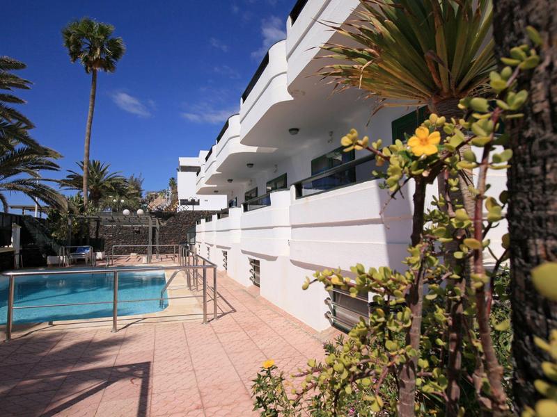 Otoczony palmami basen na zewnątrz przy białym budynku pod jasnym niebem.