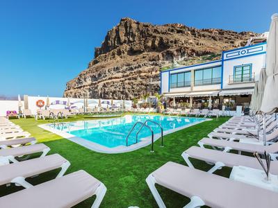 Poolbereich mit Liegestühlen und Restaurant vor felsigem Berg unter blauem Himmel.