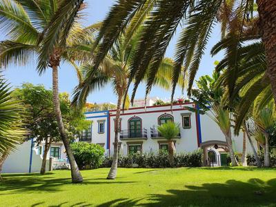 Hotelgebäude mit weißen Wänden und buntem Dach hinter Palmen und Rasenfläche bei blauem Himmel.
