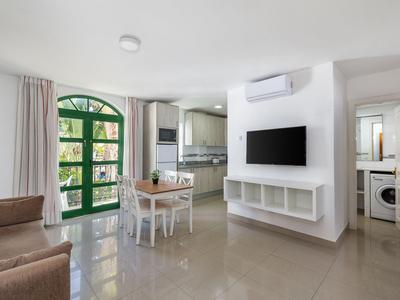 Helles Wohnzimmer mit Esstisch, Fernseher an der Wand und Sicht zur Küche und Waschraum.