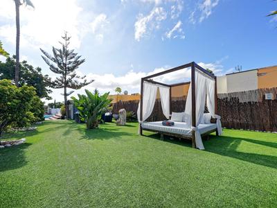 Een tuin met gras, een wit overdekt ligbed, omringd door bomen en gebouwen