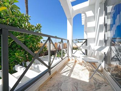 Balkon mit Metallgeländer, weißen Möbeln und Pflanzen bei sonnigem, blauem Himmel.