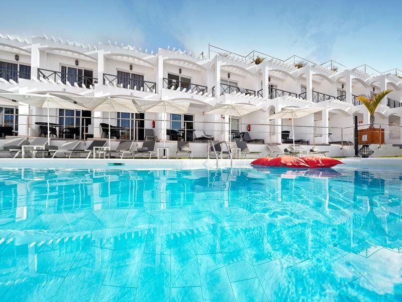 Großer blauer Pool vor weißem Hotelgebäude mit Terrasse und Liegestühlen bei klarem Himmel.