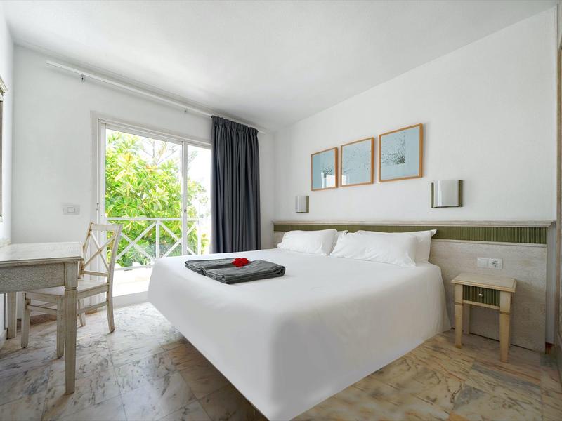 Helles Schlafzimmer mit Doppelbett, Holzmöbeln, großen Fenstern und Balkon mit grünem Blick.