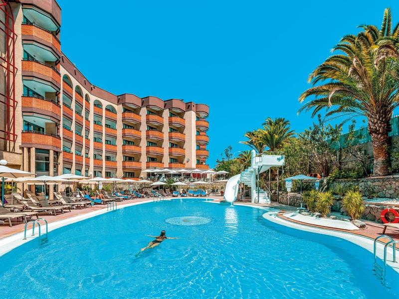 Hotel curvo con grande piscina, scivolo, palme e lettini sotto un cielo limpido.