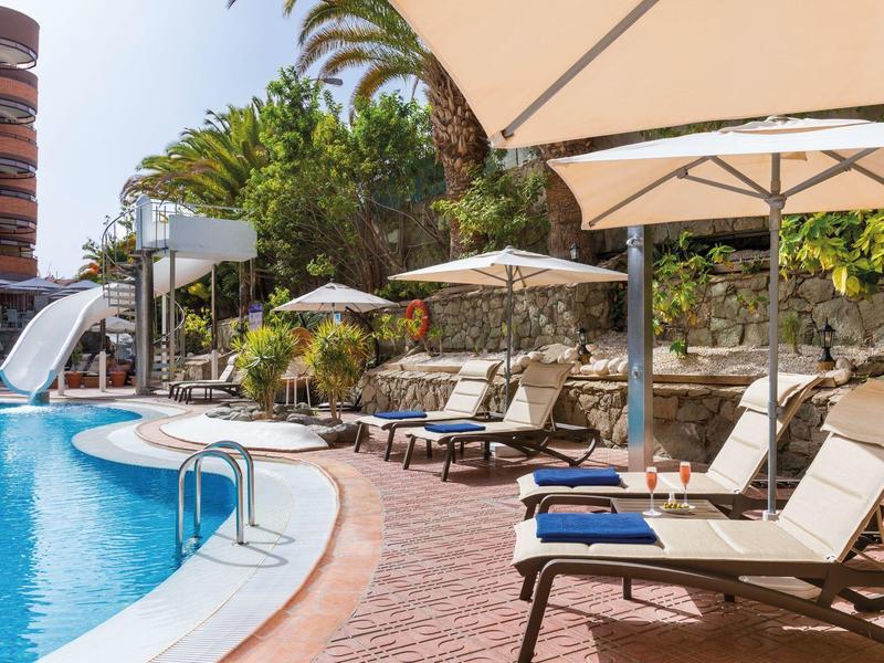 Bordo piscina con lettini e ombrelloni in un ambiente tropicale soleggiato.