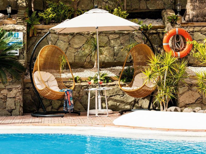 Accogliente zona relax a bordo piscina con due sedie in vimini e un ombrellone bianco accanto a un muro di pietra.