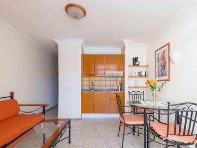 Jasný obývací pokoj s oranžovými polštáři, skleněným stolem a malou kuchyní v pozadí.